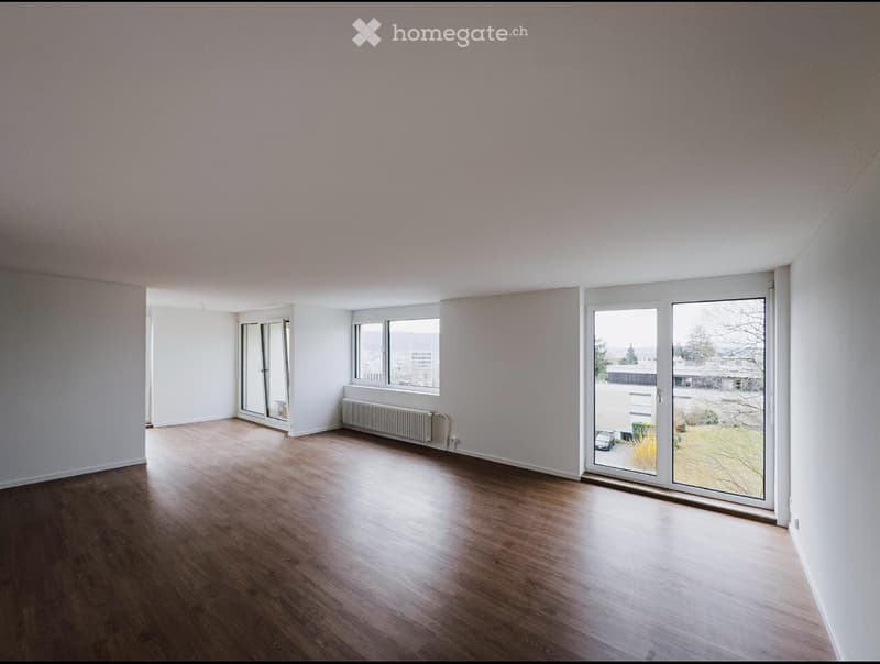 Empty room with wooden flooring, white walls, large windows, and sliding glass doors leading to an outdoor area.