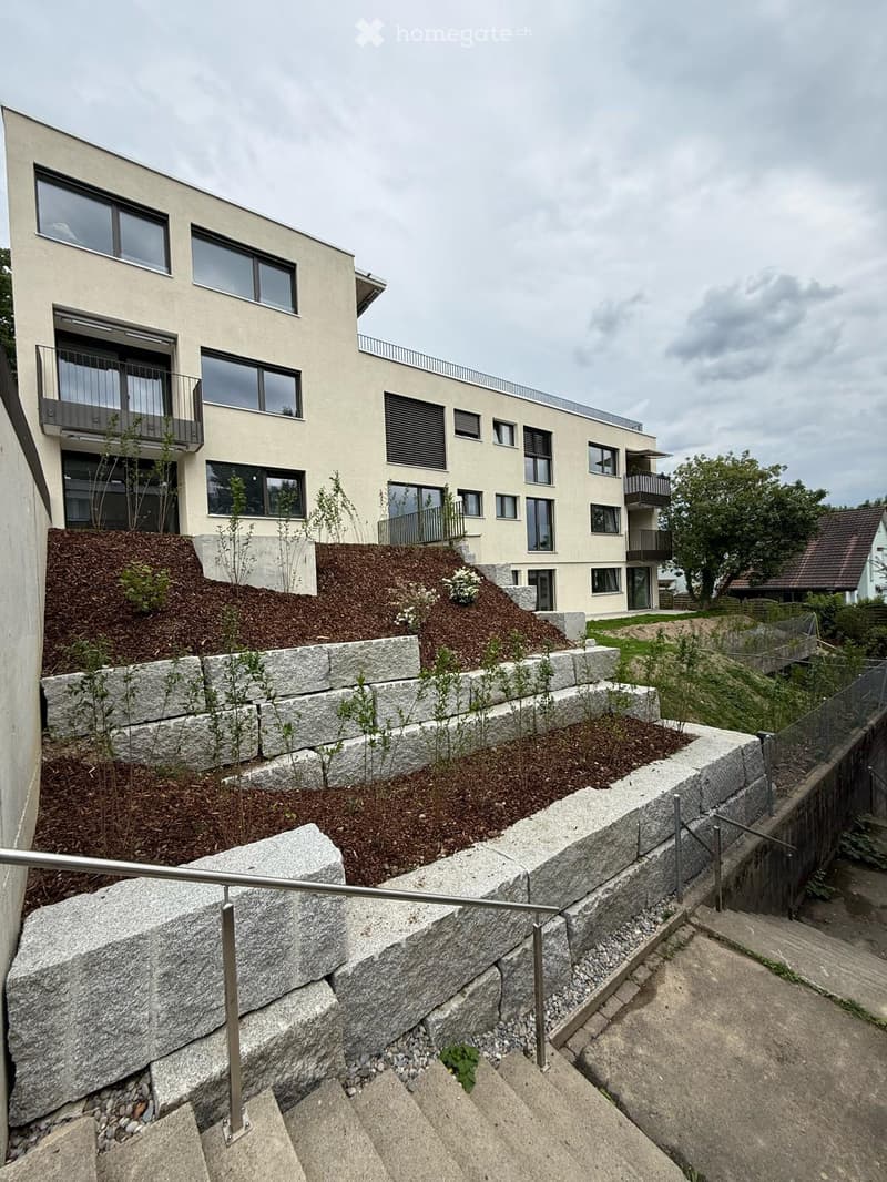 Modern multi-story building with large windows and balconies, surrounded by landscaped garden with stone walls and small plants.