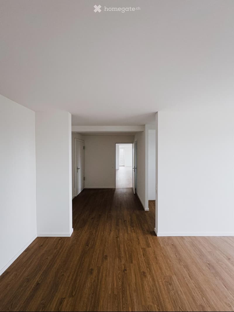 Empty hallway with wooden flooring and white walls, featuring doors at both ends and a visible room beyond.