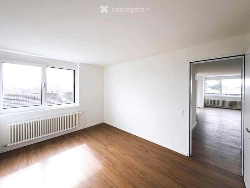 Empty room with wooden flooring, white walls, and large windows offering a view of rooftops. Radiators are visible along the walls.