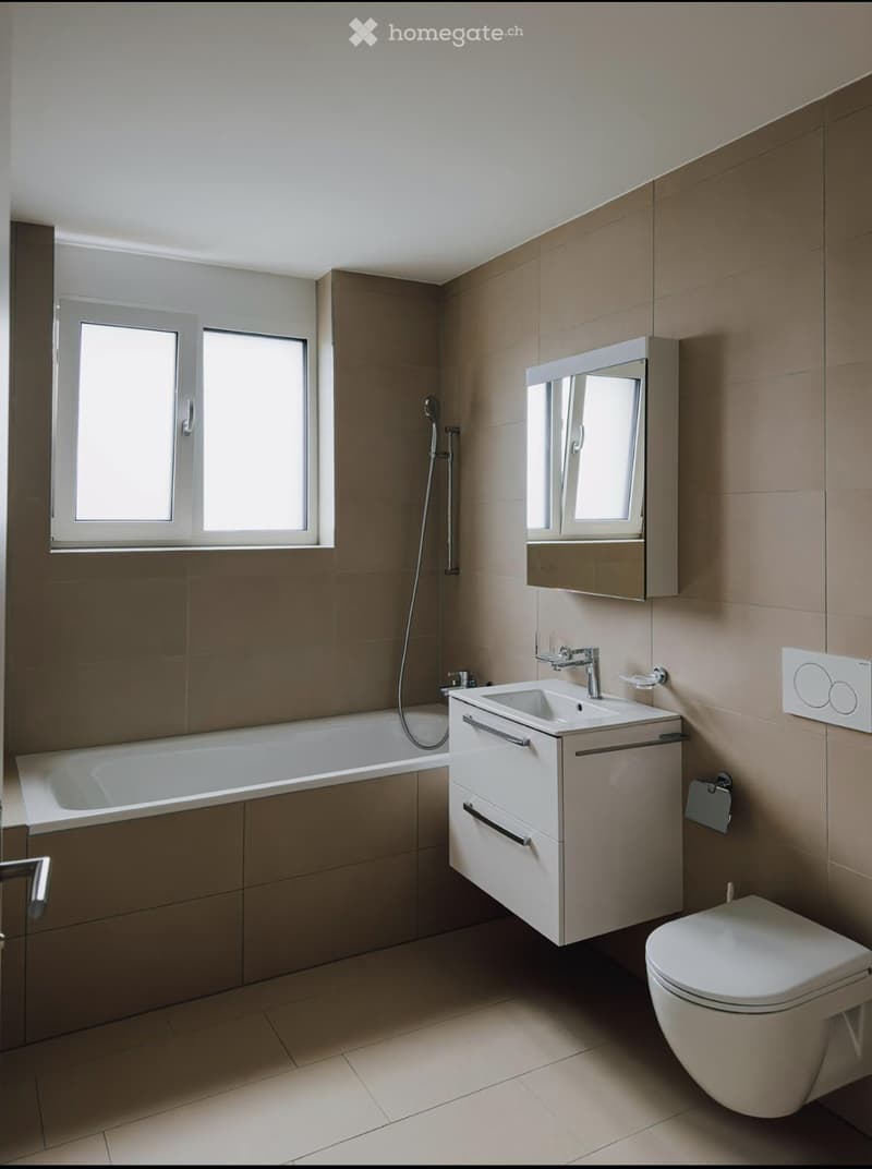 Modern bathroom with beige tiles, a bathtub, a sink with a cabinet, a toilet, and a mirrored cabinet above the sink.