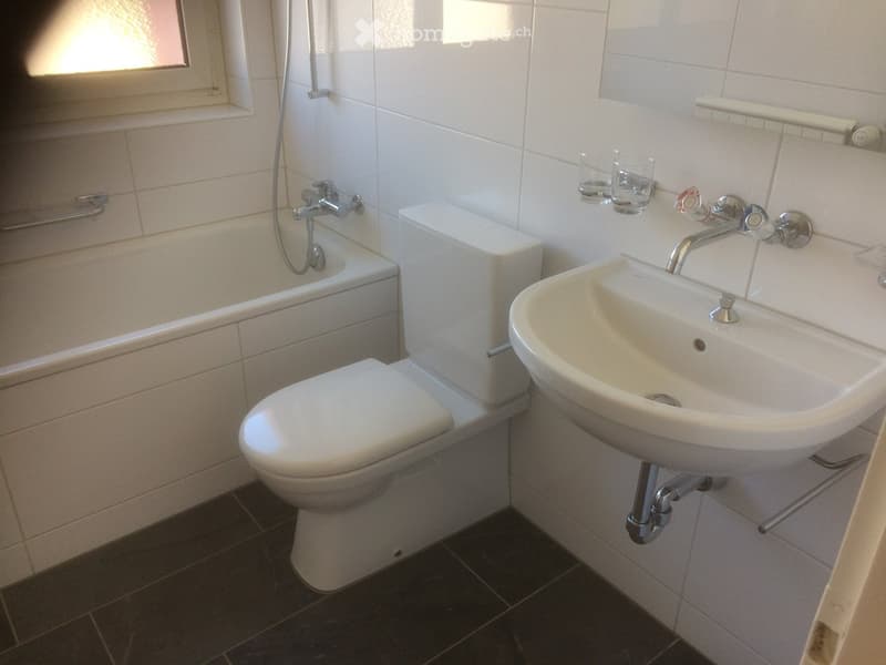 Modern bathroom with white tiles, a toilet, a sink with a chrome faucet, a bathtub with a handheld shower, and two glass cups on a shelf.