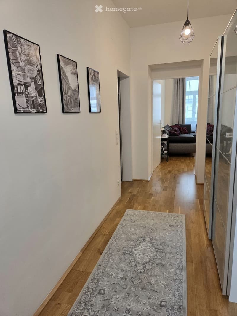 A well-lit hallway with wooden flooring, a patterned rug, and three framed black-and-white pictures on the wall. The hallway leads to a living room area with a sofa and side table visible in the background.