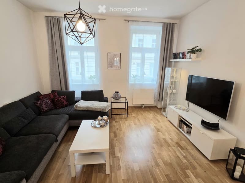 Modern living room with wooden flooring, large windows, grey sectional sofa, white coffee table, TV stand, side table, wall shelves, and a geometric hanging light fixture.