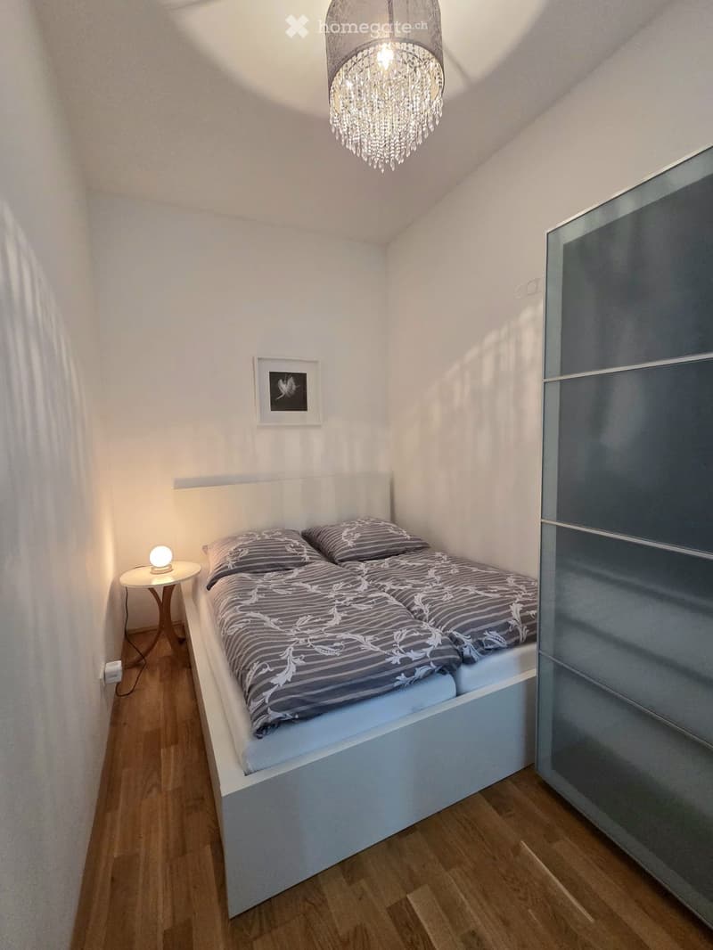 Cozy bedroom with a white bed, grey bedding, a small nightstand with a lamp, a modern wardrobe, and a decorative picture frame on the wall.