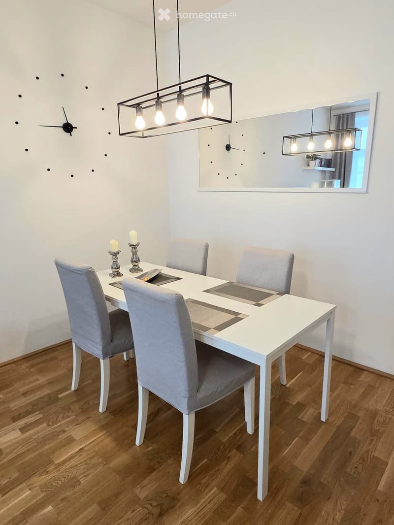 Dining room with a white dining table, four grey upholstered chairs, two candles on the table, and modern light fixtures.