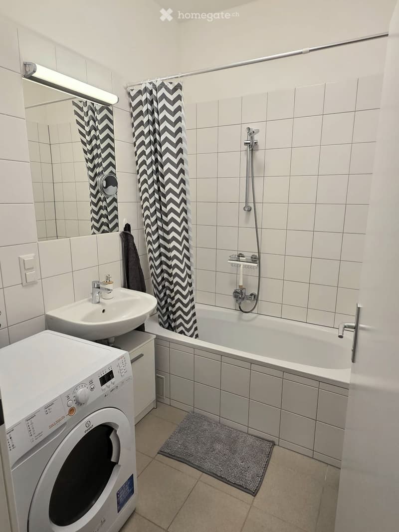 Modern bathroom with white tiles, a sink, bathtub with shower, washing machine, and a towel on the wall.