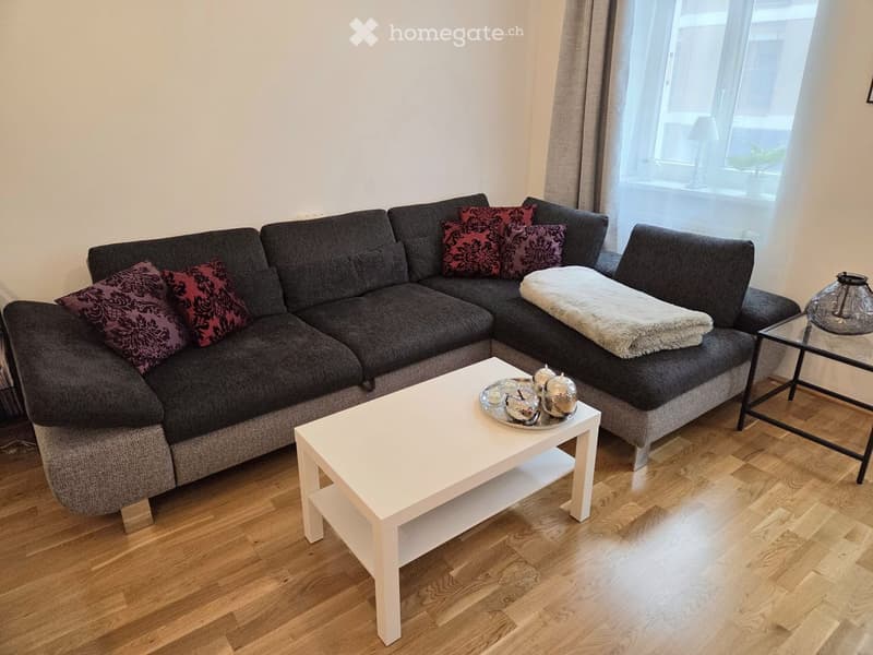 Modern living room with a grey sectional sofa, white coffee table, decorative tray with candles, and a glass table lamp. The room features wooden flooring and large windows with sheer curtains.