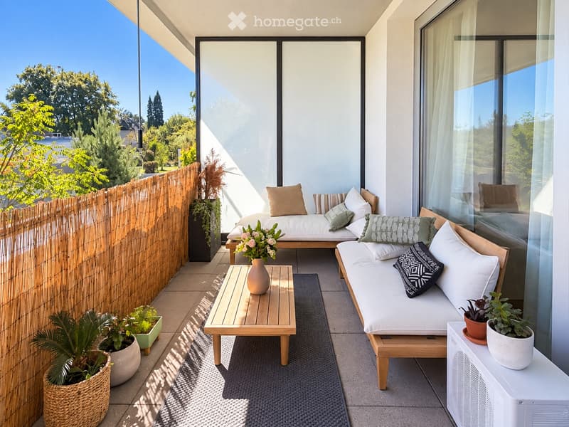 Modern balcony with a wooden sofa, small wooden table, vase with flowers, and several potted plants. The space features a bamboo privacy screen and large windows overlooking a green area.