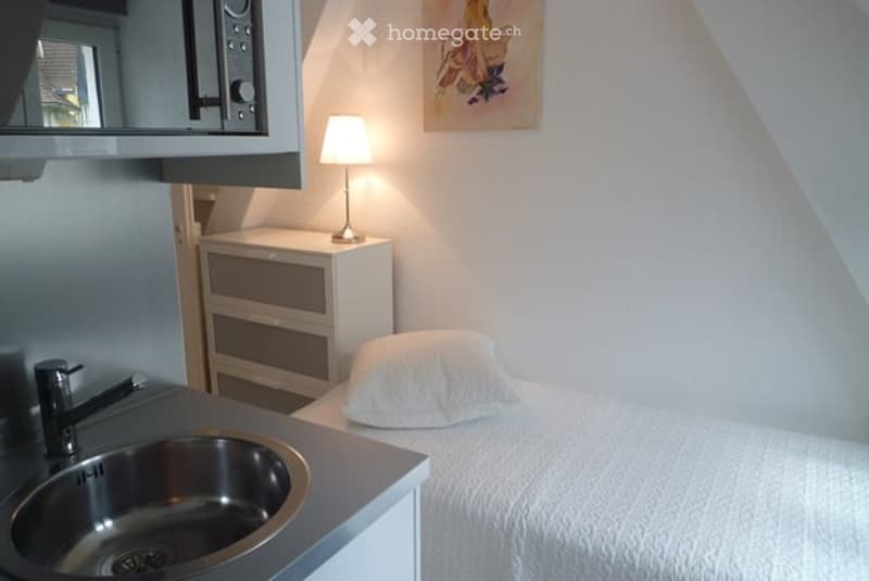 Cozy bedroom with a single bed, nightstand with a lamp, a small sink with a faucet, and a mirror above the nightstand. The room features white walls and a slanted ceiling typical of an attic space.