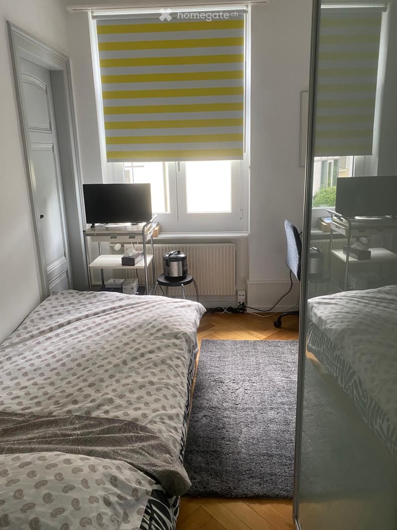 A small bedroom with two single beds, a small table with a screen, a chair, a stool with a rice cooker, and a radiator under the window. The room has wooden flooring and a grey rug.