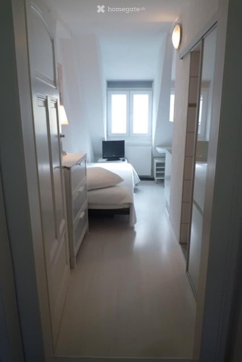 A small, modern bedroom with a bed, nightstand, lamp, and a mirror. The room features light-colored walls and wooden flooring.