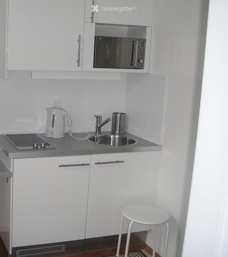 Compact kitchen with white cabinets, microwave above counter, electric kettle, sink with faucet, and a white stool.