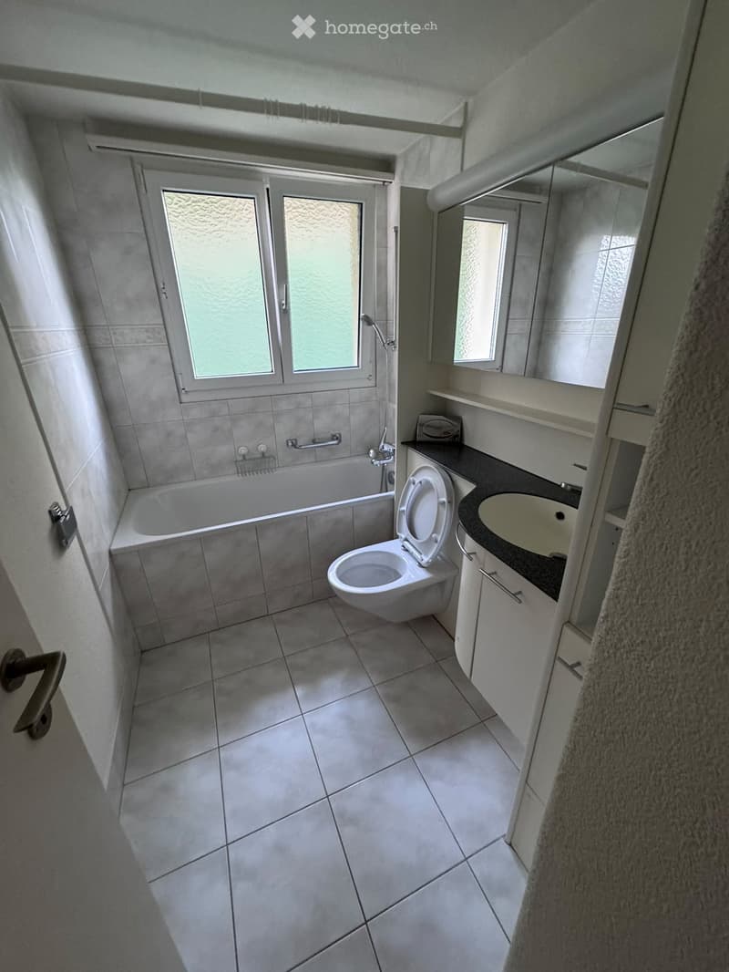 Modern bathroom with tiled walls and floor, featuring a bathtub, toilet, sink, mirror, and cabinet.