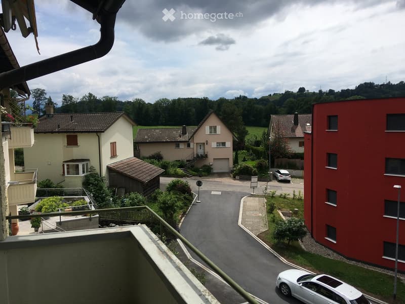 Balcony view overlooking a quiet residential street with houses, greenery, and parked cars.