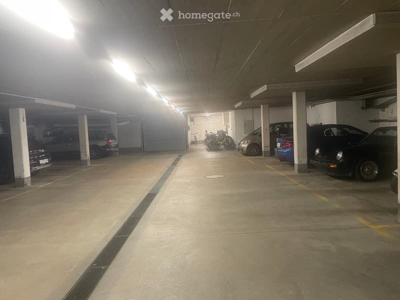 Underground parking area with several parked cars and bicycles, concrete floor, fluorescent lighting, and support pillars.