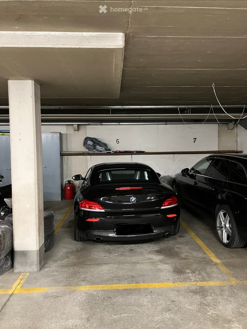 Underground parking area with two parked cars, a BMW Z4 and another sedan, marked parking spaces, and a fire extinguisher on a shelf.