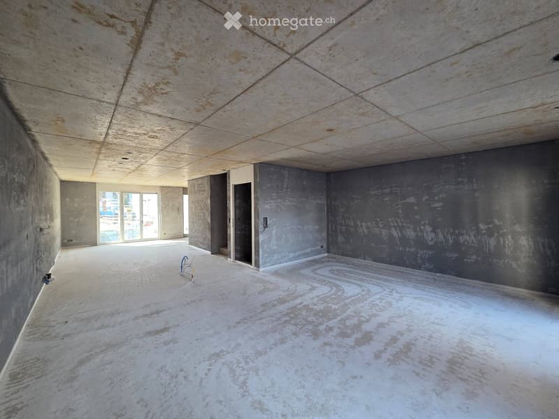 Empty room with bare concrete walls and ceiling, unfinished surfaces, and a large window letting in natural light.