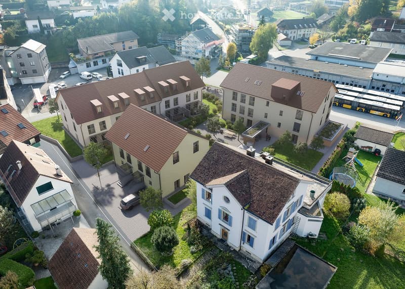 Aerial view of a residential area with multiple houses, green spaces, and a parking lot. The houses have brown tiled roofs and are surrounded by trees and lawns.