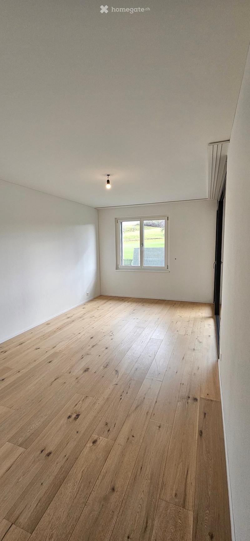 Empty room with light wooden flooring, white walls, and a single window with a view of a green outdoor area.