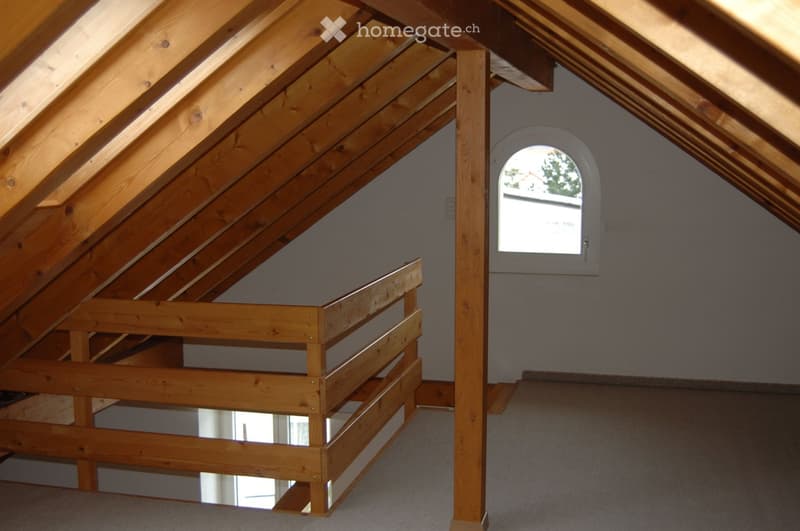 An empty attic space with wooden beams and a small window, featuring exposed wooden construction and a carpeted floor.
