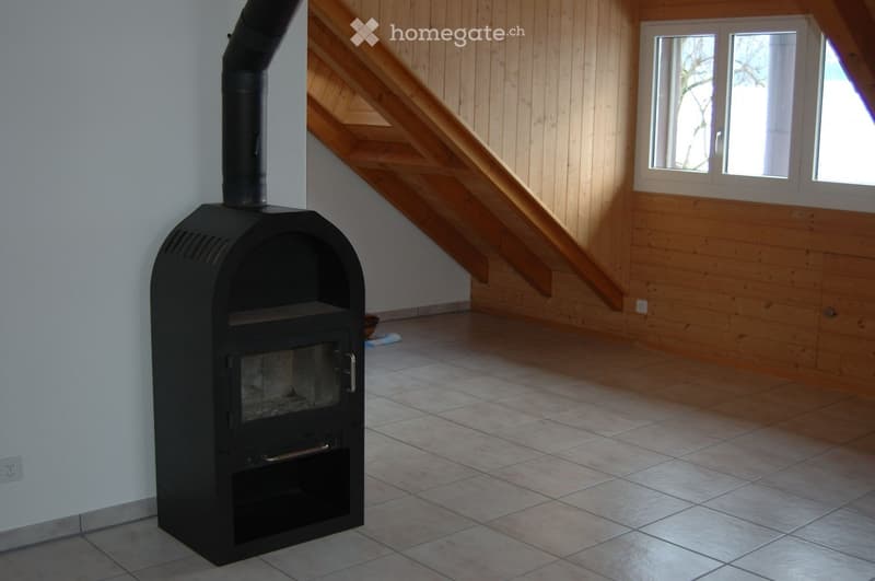 Empty room with wooden walls, tiled floor, and a black wood-burning stove with a chimney. A wooden staircase leads upwards, and a window with white frames is visible on the right side.