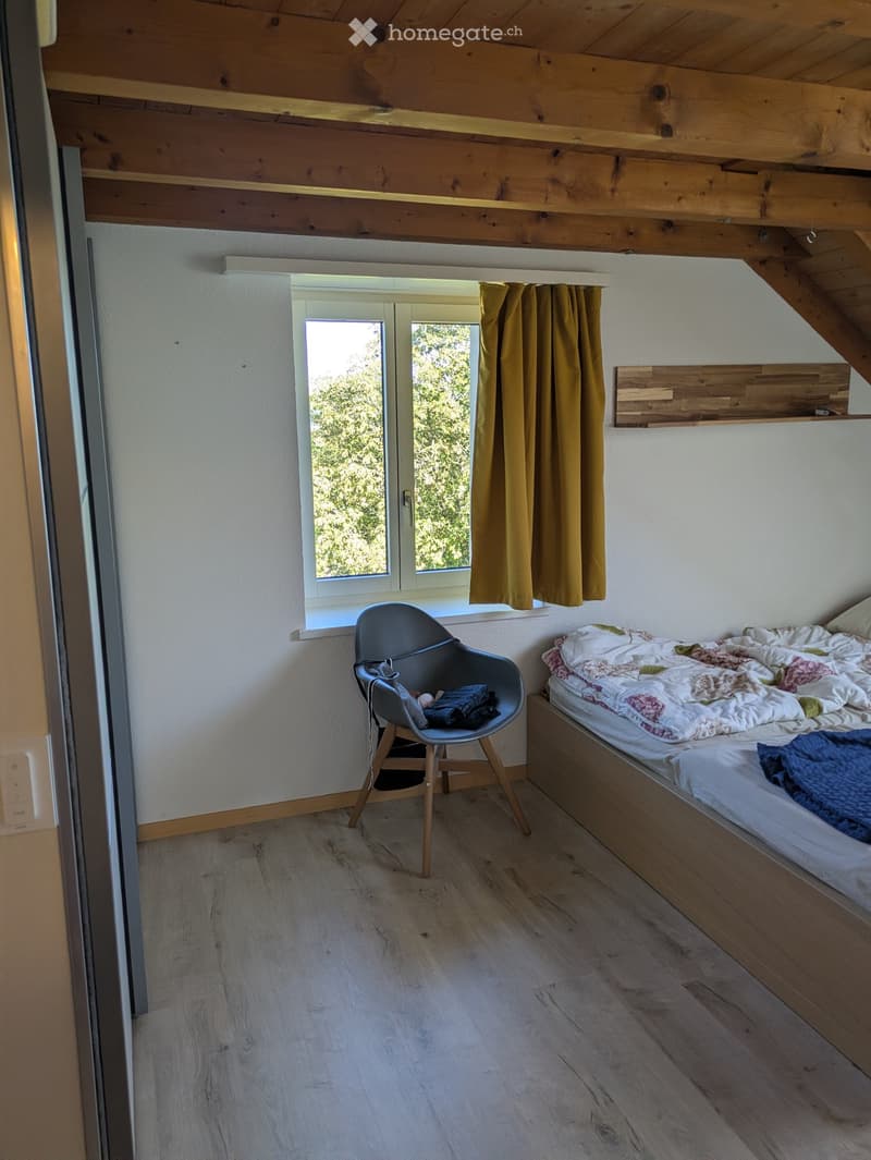Cozy bedroom with wooden floors, slanted wooden ceiling beams, a bed with a colorful quilt, a modern chair, and a window with yellow curtains.
