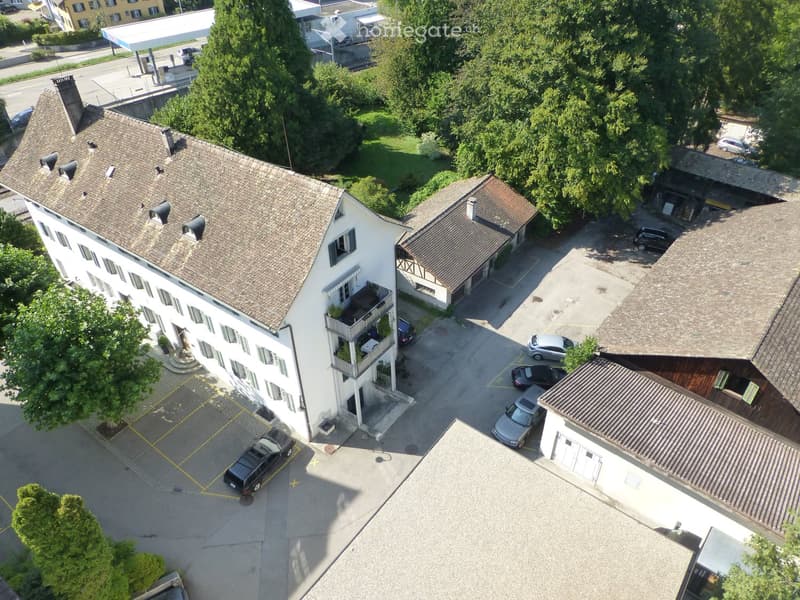 Aerial view of a multi-story residential building with a tiled roof, surrounded by trees and parked cars in a paved area.