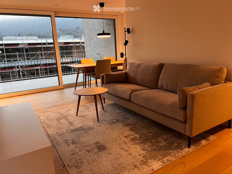 Modern living room with a brown sofa, coffee table, dining table, and chairs, large windows with a view, and wooden flooring.