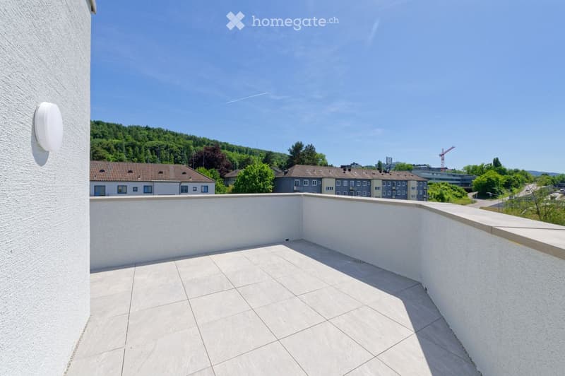 A clean, tiled balcony with white walls and a low railing, overlooking a residential area with houses, trees, and a clear blue sky.