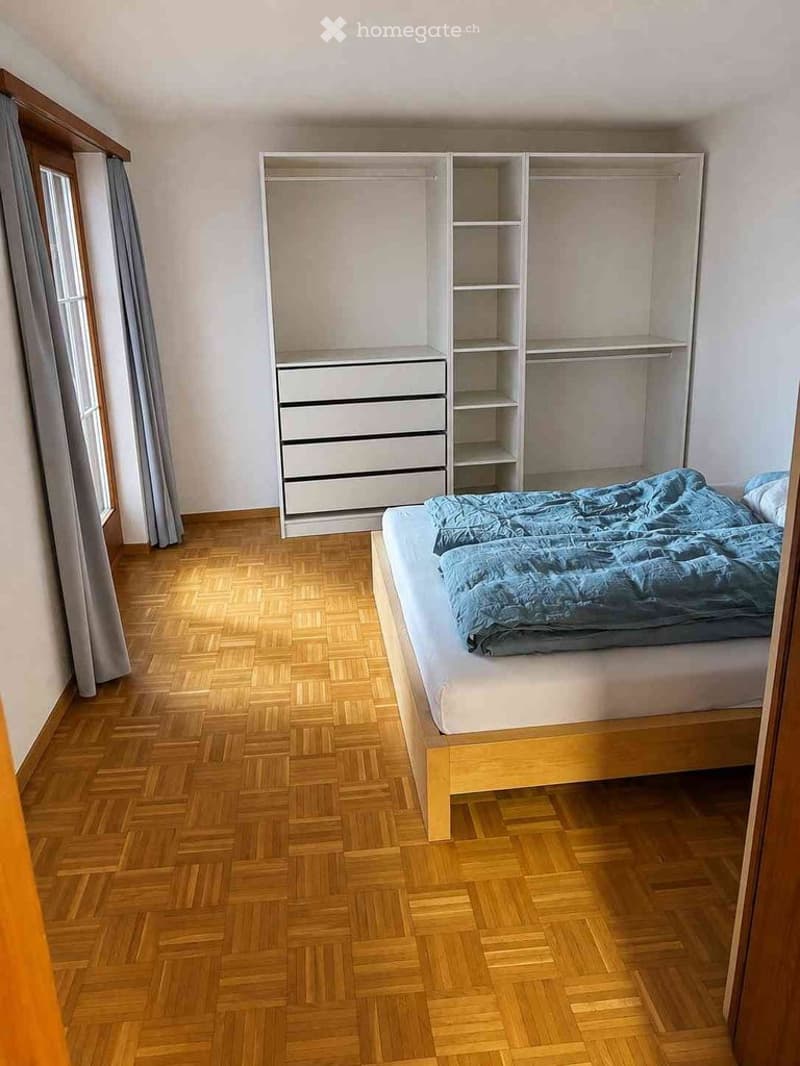 Bedroom with wooden herringbone flooring, a single bed with blue bedding, a built-in wardrobe with shelves and drawers, and a window with grey curtains.