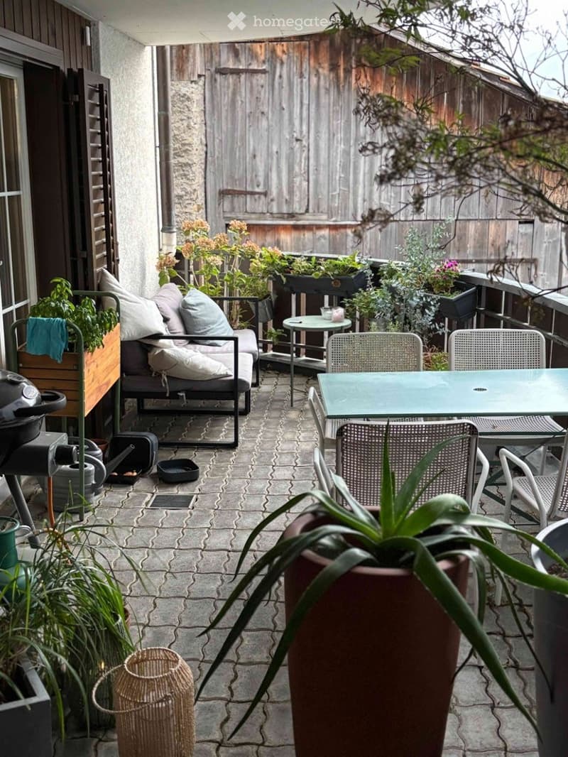 A cozy outdoor balcony with a seating area, small table, chairs, and various potted plants. The space features a brick floor and wooden fencing.