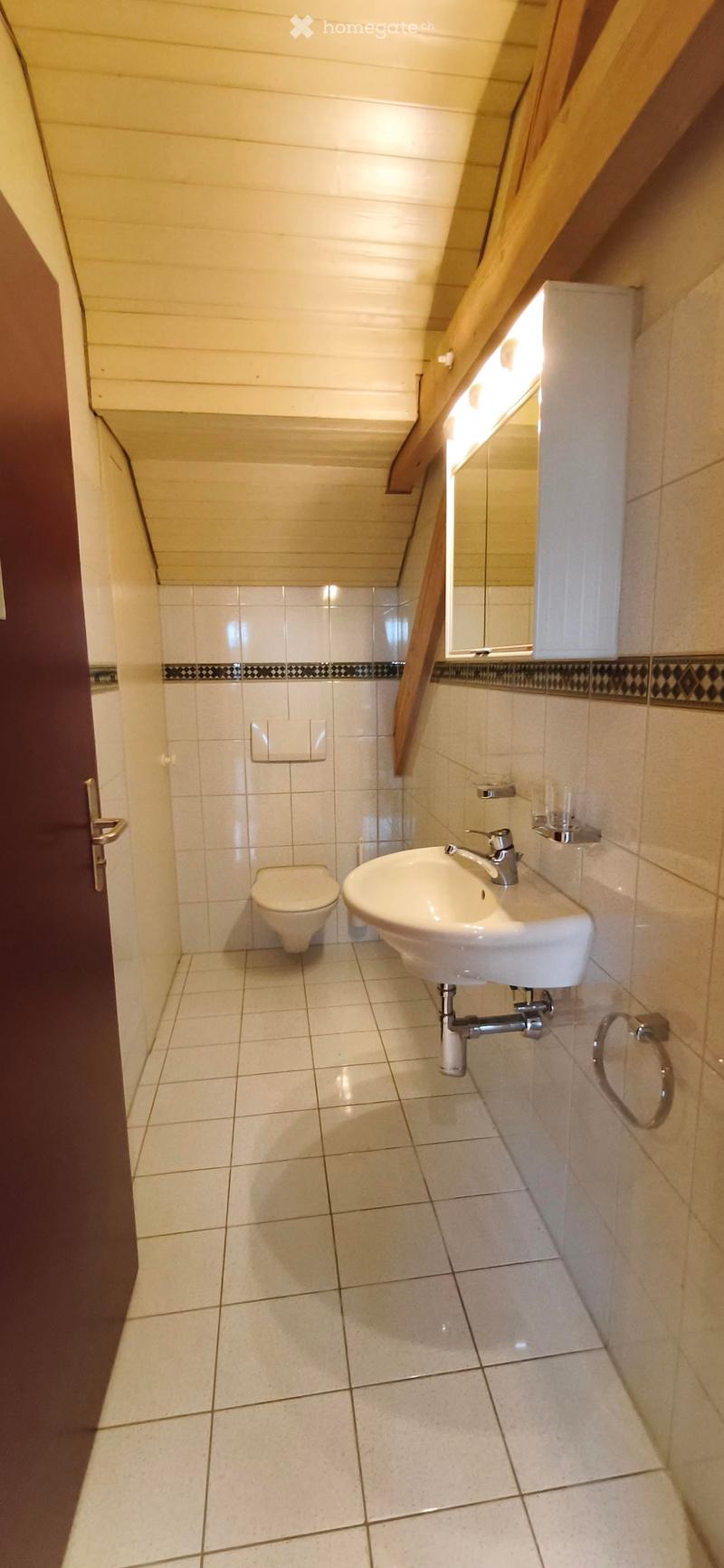 A small, clean bathroom with white tiled walls and floor, featuring a toilet, a sink with a modern faucet, a mirror above the sink, and a towel ring on the wall.