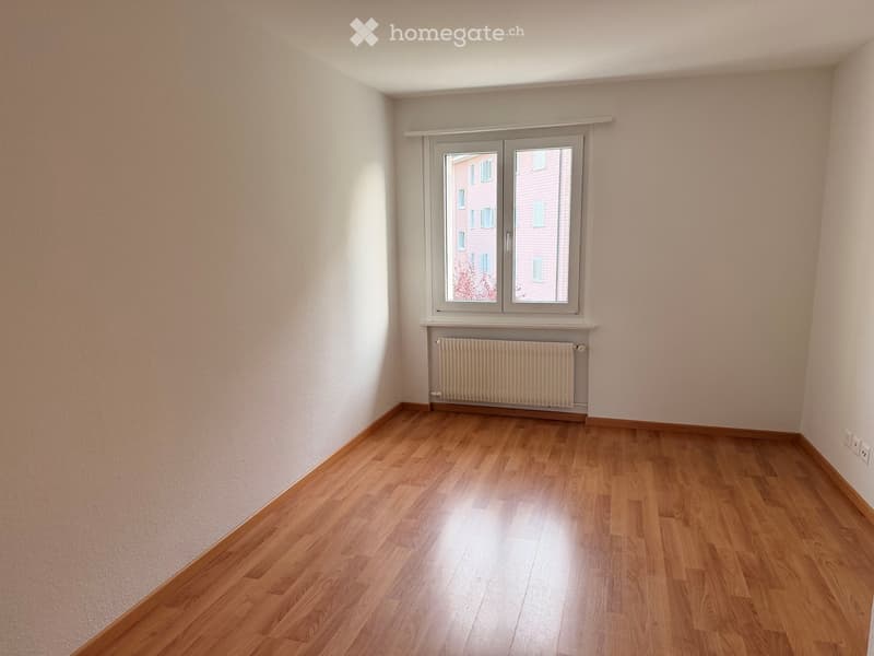 Empty room with wooden flooring, white walls, a window with a radiator below, and a visible electrical outlet on the wall.