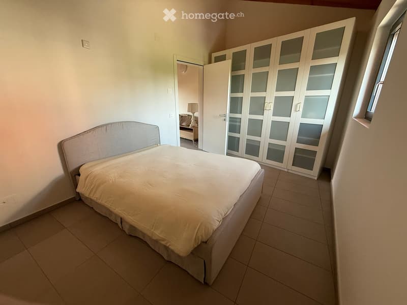 A well-lit bedroom with a neatly made bed, a large wardrobe with glass doors, and a small lamp visible in the adjacent room.