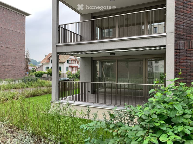 Modern balcony with metal railing, glass doors, and a view of a green garden and neighboring houses.