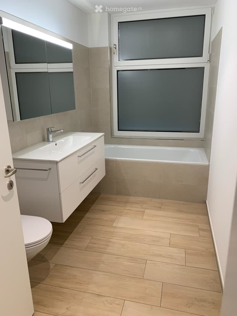 Modern bathroom with a white sink, chrome faucet, bathtub, toilet, and large mirror. Light wood flooring and neutral-colored tiles.