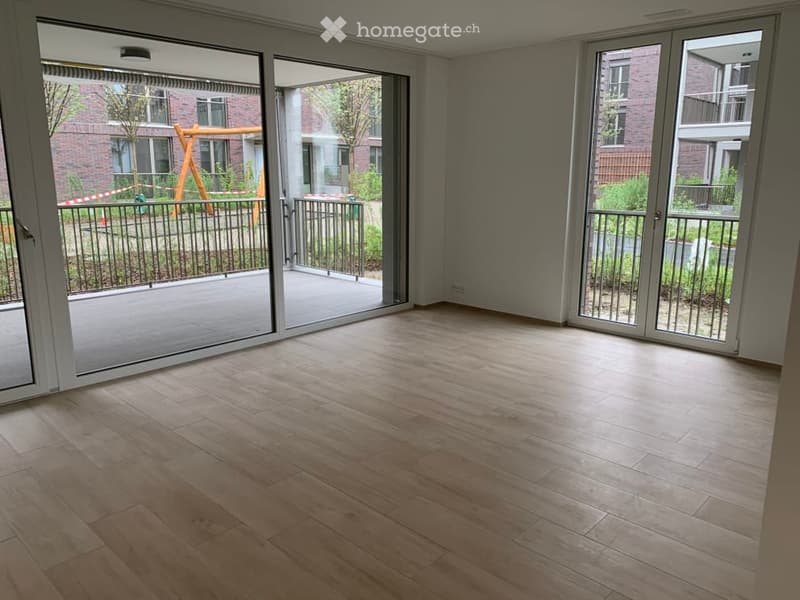 Empty room with wooden flooring, large sliding glass doors leading to a balcony, and a view of a playground and neighboring buildings.