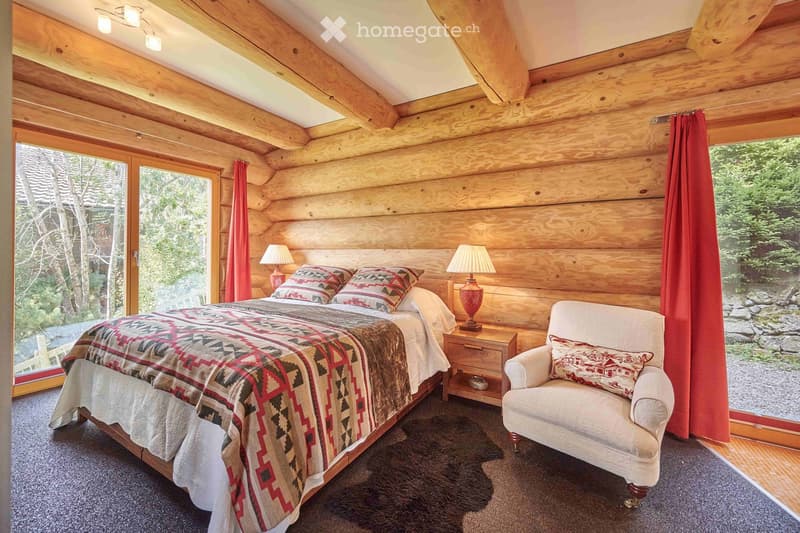 log cabin bedroom with wooden bed and white chair, night table with lamps, blanket and pillows, curtains, rug and windows with tree view