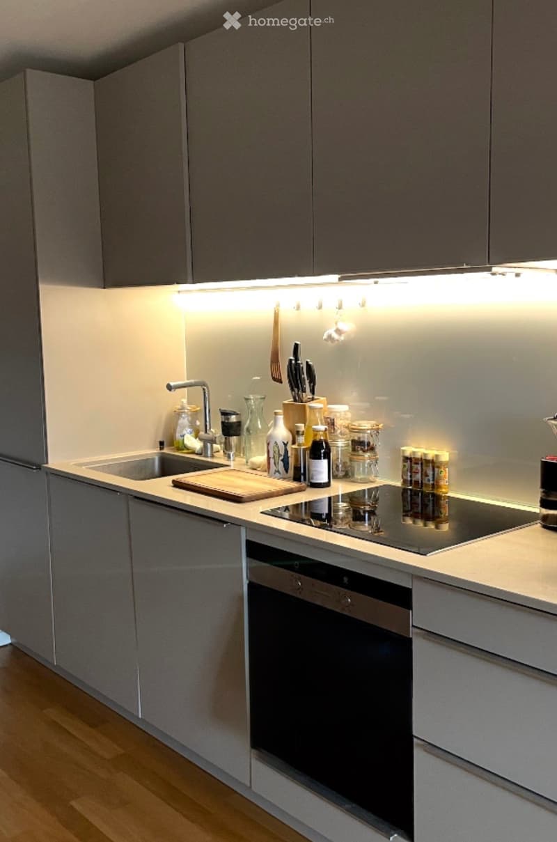 Modern kitchen with grey cabinets, stainless steel appliances, and a sleek countertop. Features include a sink, faucet, cutting board, blender, oven, and various kitchen items on the counter.