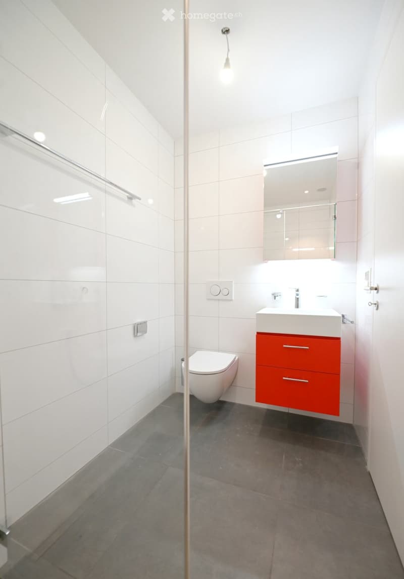 Modern bathroom with white tiles, a toilet, a sink with a red cabinet, and a large mirror above the sink.