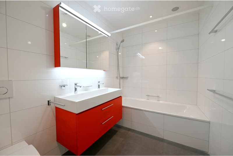 Modern bathroom with white tiles, red cabinetry, a large sink, bathtub, and a mirror with built-in lighting.