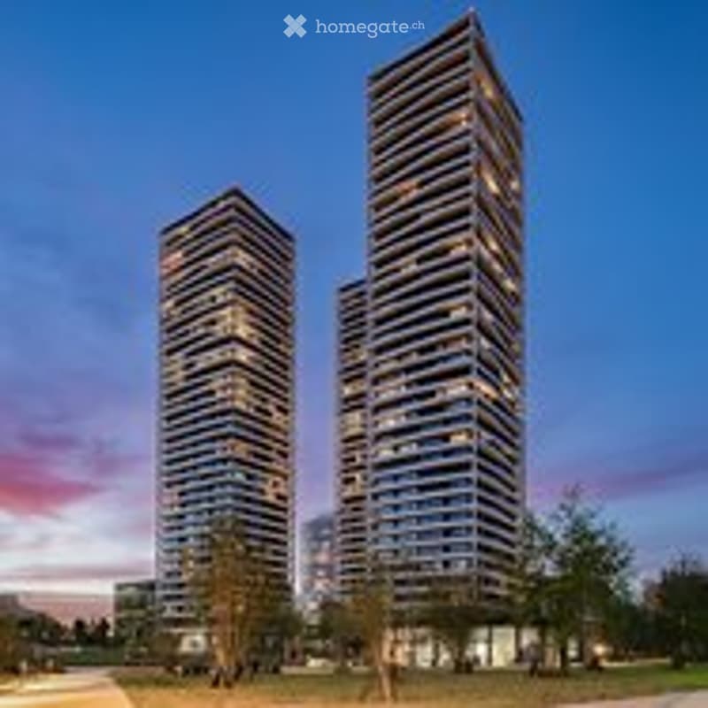 Two modern high-rise residential buildings with glass facades, set against a twilight sky with pink and purple hues. The buildings have multiple floors and balconies, surrounded by a landscaped area with trees and open space.