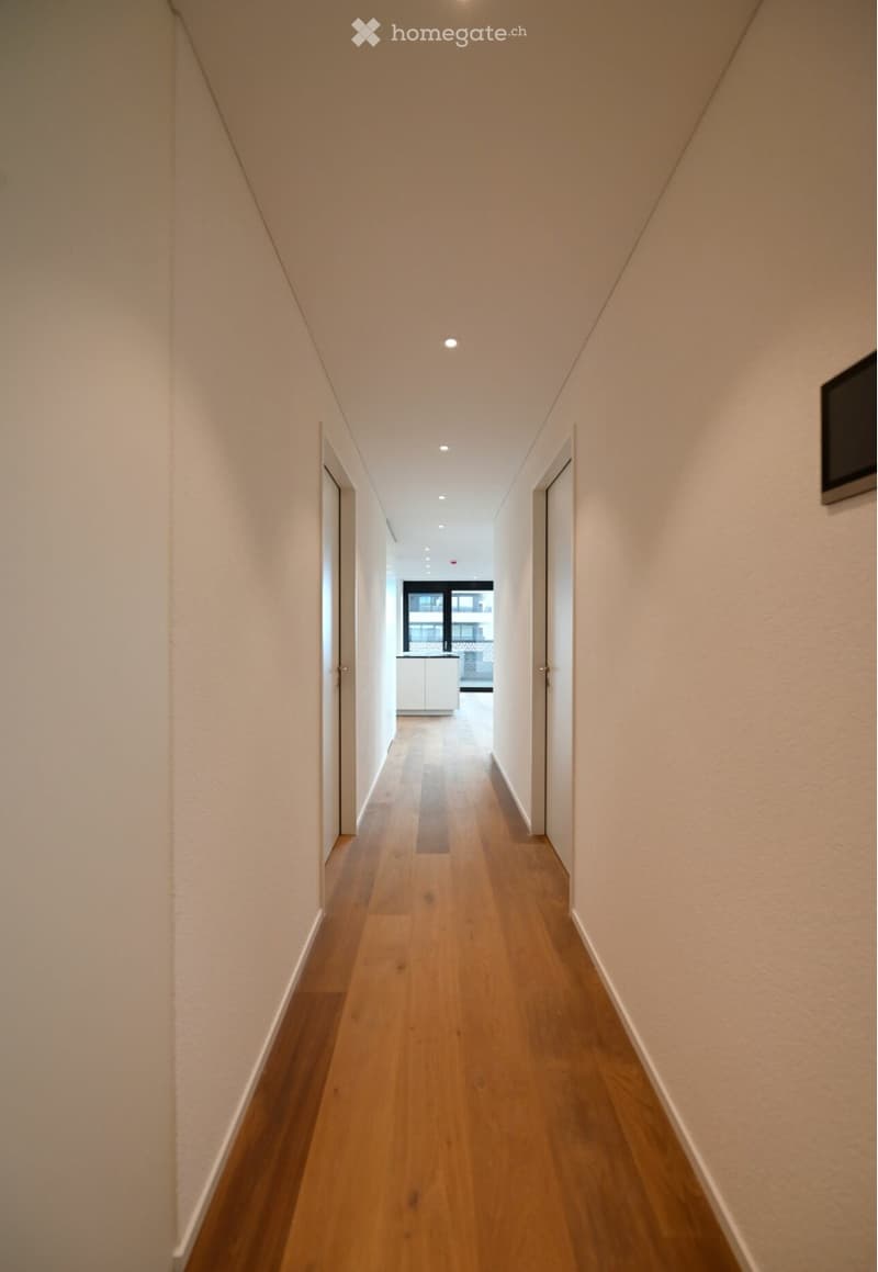 A narrow hallway with light wooden flooring, white walls, and recessed ceiling lights. Doors are visible on both sides, leading to other rooms.
