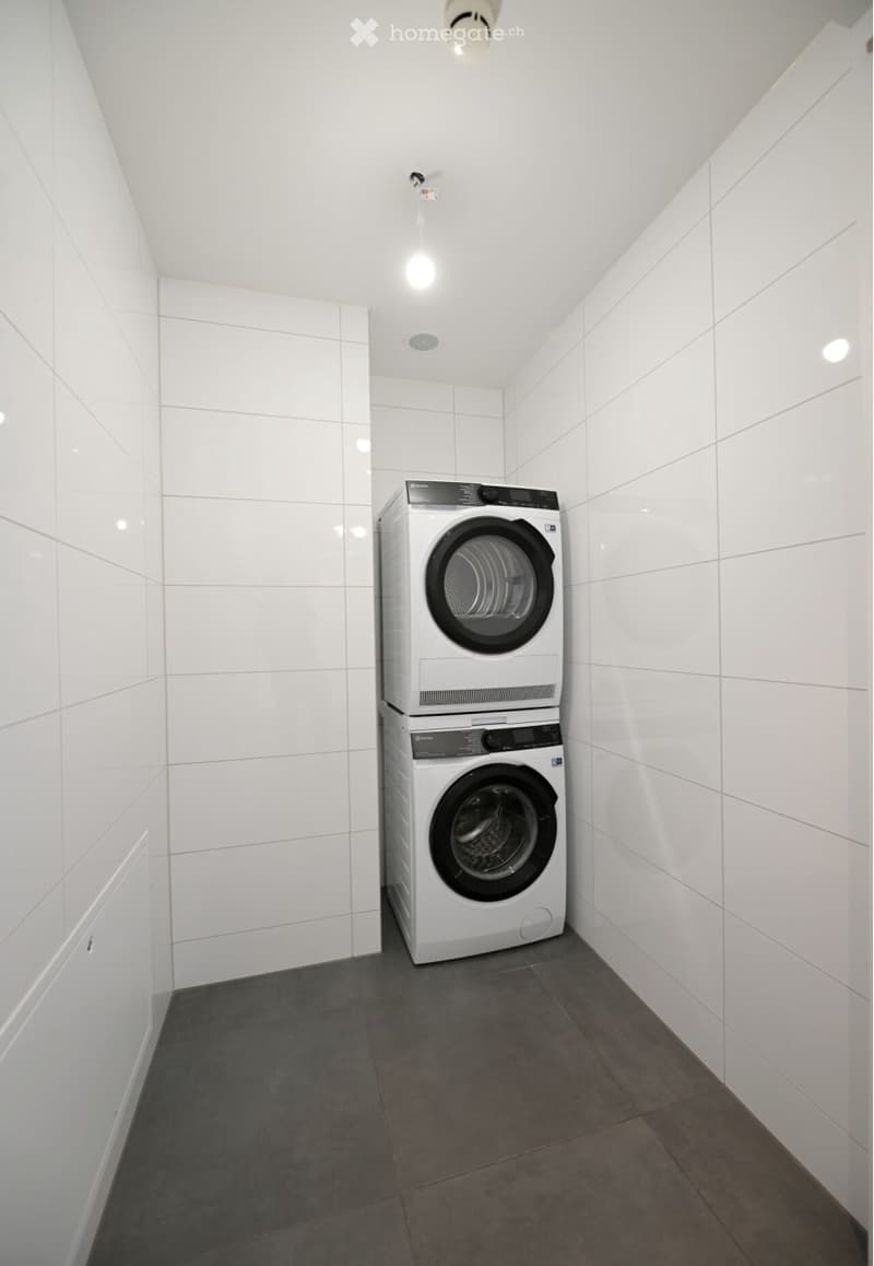 Modern, compact laundry room with white tiled walls and dark grey tiled floor, featuring a stacked washer and dryer unit.