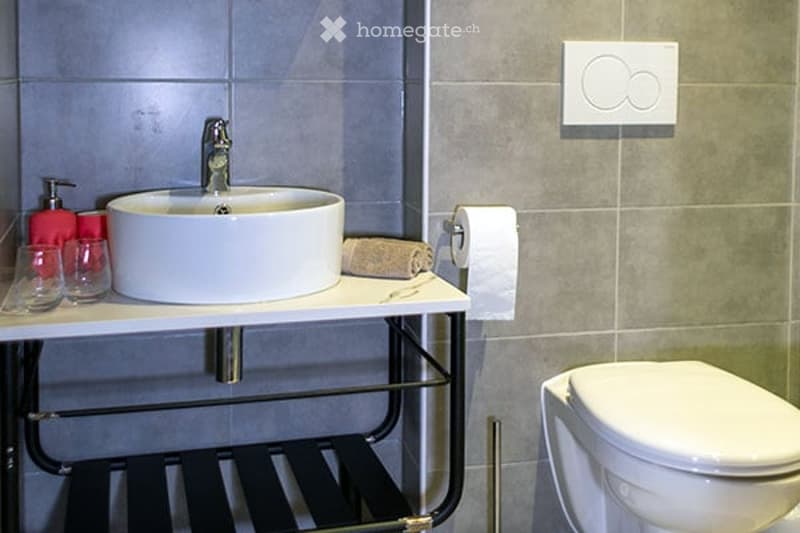 Modern bathroom with a white sink, chrome faucet, toilet, and a towel on the counter. There are two glasses and a soap dispenser on the counter as well.