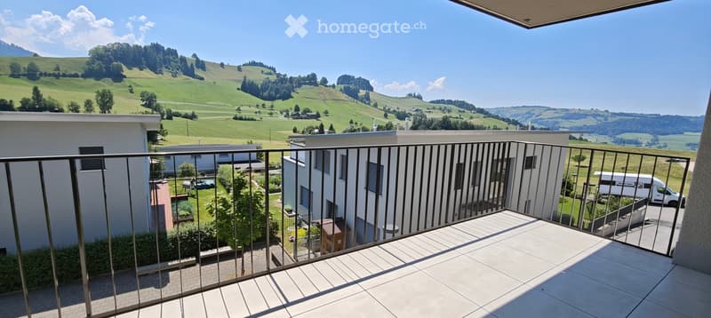 Balcony with metal railing overlooking a green, hilly landscape with scattered trees and buildings below.