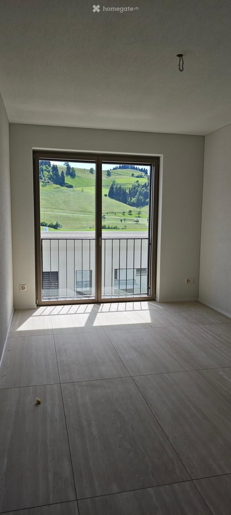 Empty room with large sliding glass doors leading to a balcony, offering a scenic view of green hills and trees. The floor is tiled, and the walls are light-colored.