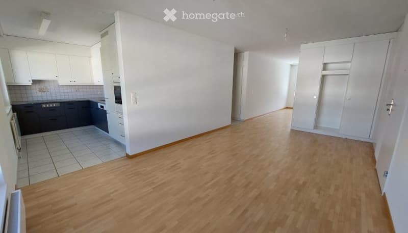 Empty room with wooden flooring, white walls, and a small kitchen area with cabinets and appliances visible in the background.