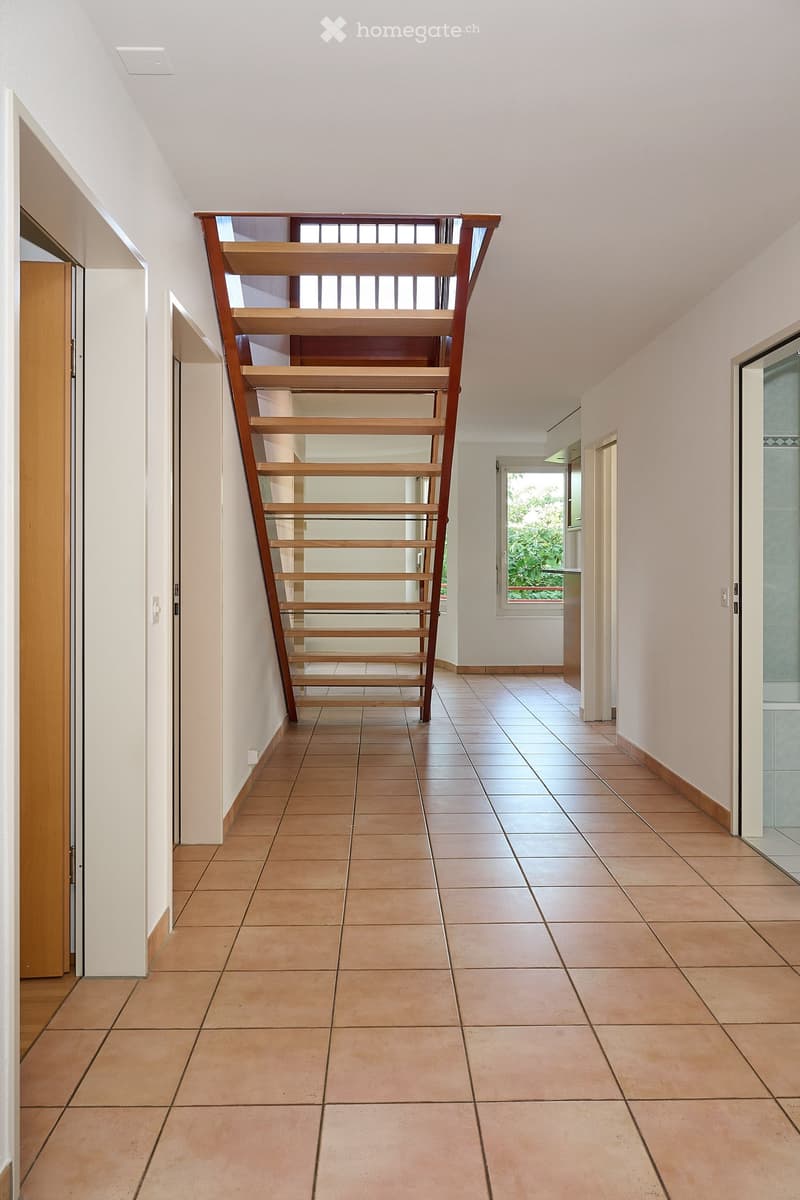 Empty hallway with tiled floor, wooden staircase, and white walls. Natural light from a window on the right side.